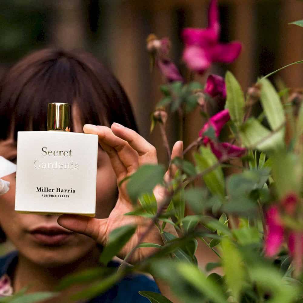 Miller Harris Secret Gardenia perfume bottle held in hand, surrounded by lush greenery and pink flowers