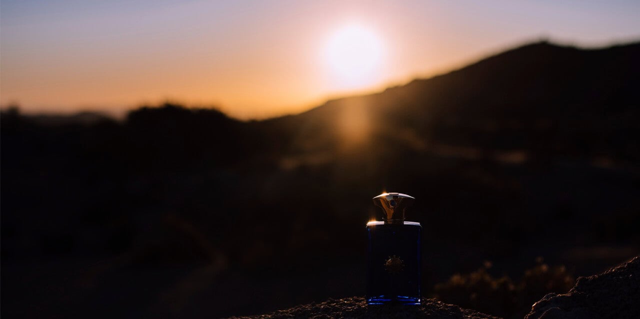 Blue perfume bottle silhouetted against a sunset mountain landscape.