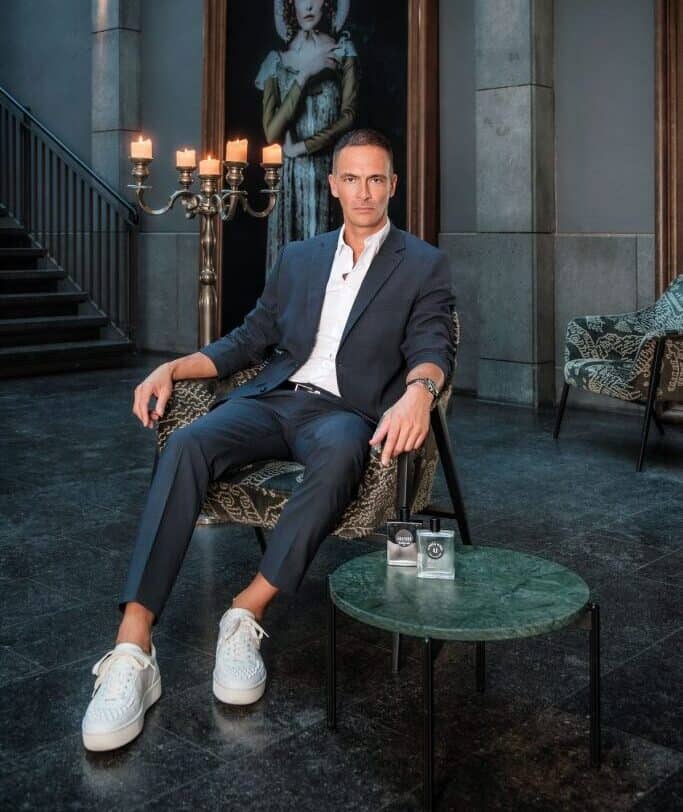Man in a suit and sneakers poses with fragrance bottles.
